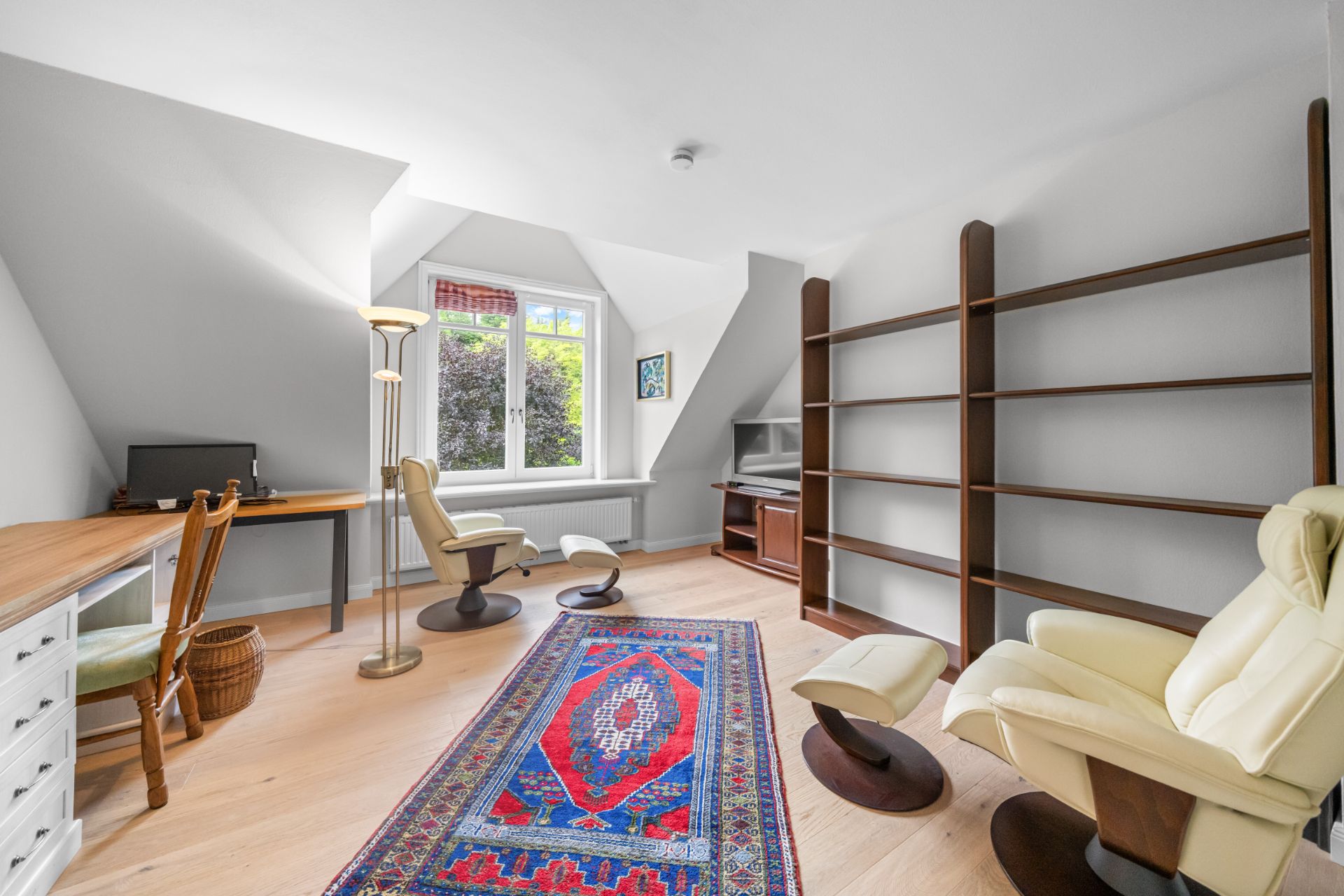 Helles Arbeitszimmer mit Teppich, Schreibtisch, Regalen, zwei Sesseln und großem Fenster.