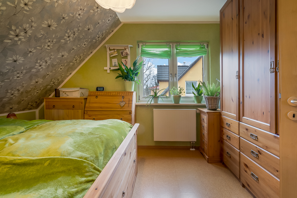 Kleines Schlafzimmer mit Holzschränken, grünem Bett und mehreren Pflanzen am Fenster.