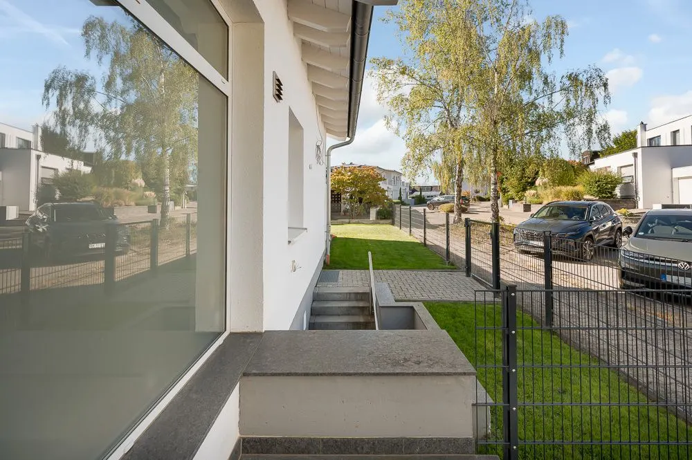 Seitenansicht eines modernen Wohnhauses mit Garten, Zaun und parkenden Autos auf der Straße.