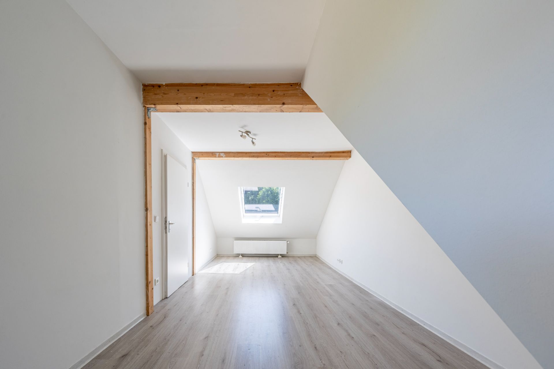 Heller Raum mit Dachschräge, Fenster, Holzdeckenbalken und hellem Boden.