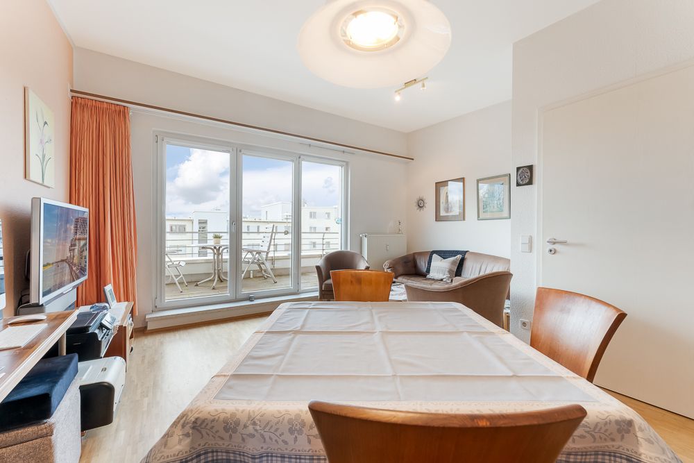 Helles Wohnzimmer mit Esstisch, Sofa, Fernseher und Zugang zu einem Balkon mit Sitzgelegenheit.