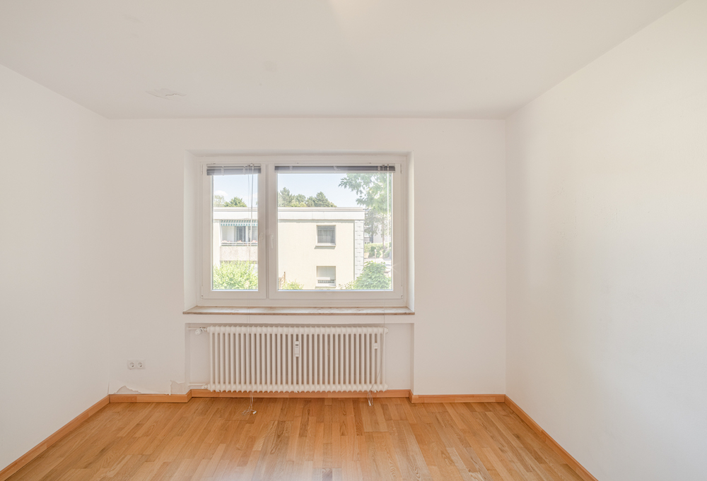 Leeres Zimmer mit Holzfußboden, weißer Wand und einem Fenster mit Blick nach draußen.