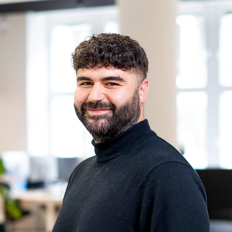 Ein Mann mit Bart und dunklem Pullover steht in einem hellen Raum mit großen Fenstern.