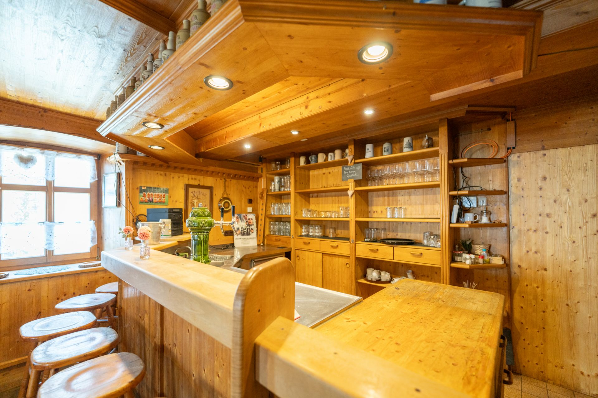 Gemütliche Wohnküche mit Holztheke, Barhockern und Regalen aus Holz, viele Gläser sichtbar.
