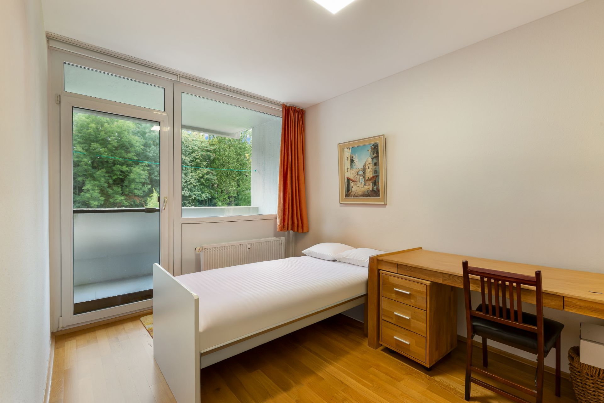 Helles Schlafzimmer mit Einzelbett, Schreibtisch, Stuhl und Zugang zu einem Balkon.