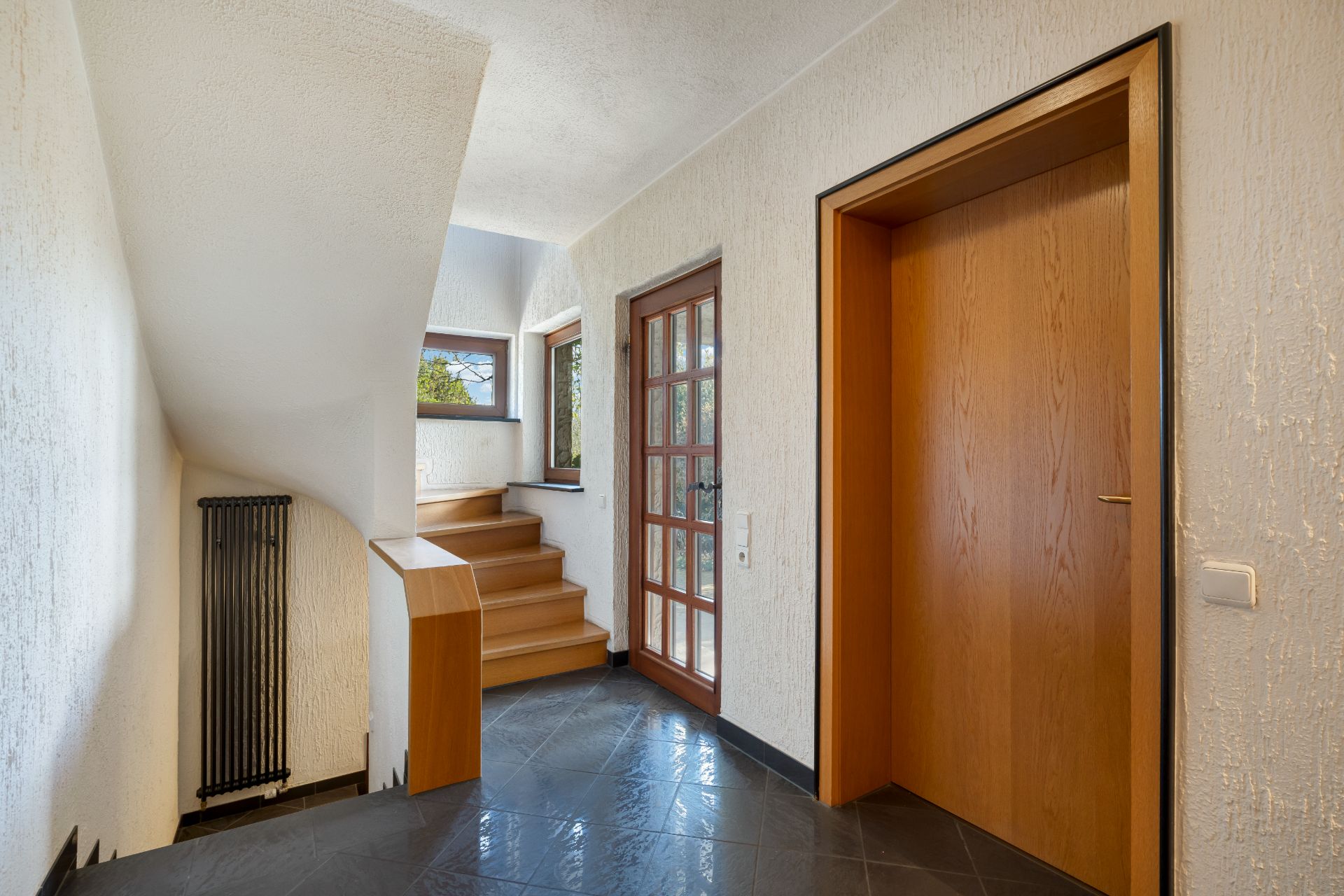 Heller Flur mit Holztreppe, Glastür, Holztür und schwarzen Fliesen.