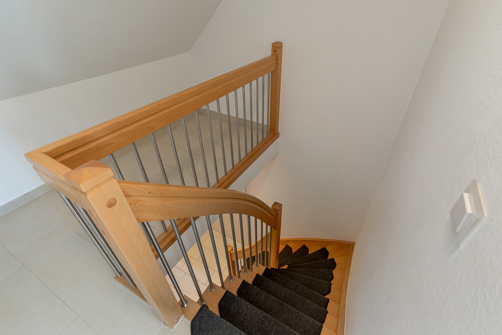 Holztreppe mit Metallgeländer in einem hellen Flur, Wand mit Lichtschalter.