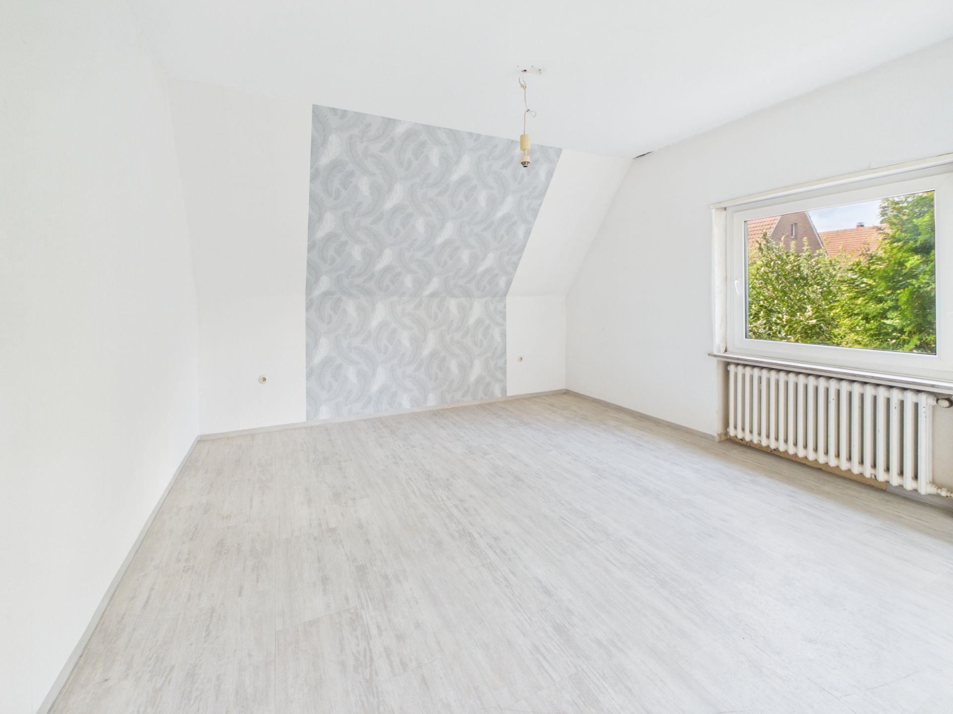 Helles, leeres Zimmer mit großem Fenster, Heizung und gemusterter Wand.