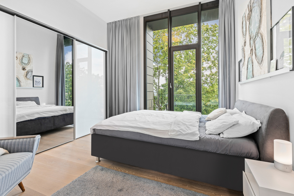 Modernes Schlafzimmer mit großem Bett, Spiegelkleiderschrank und bodentiefen Fenstern mit Blick ins Grüne.