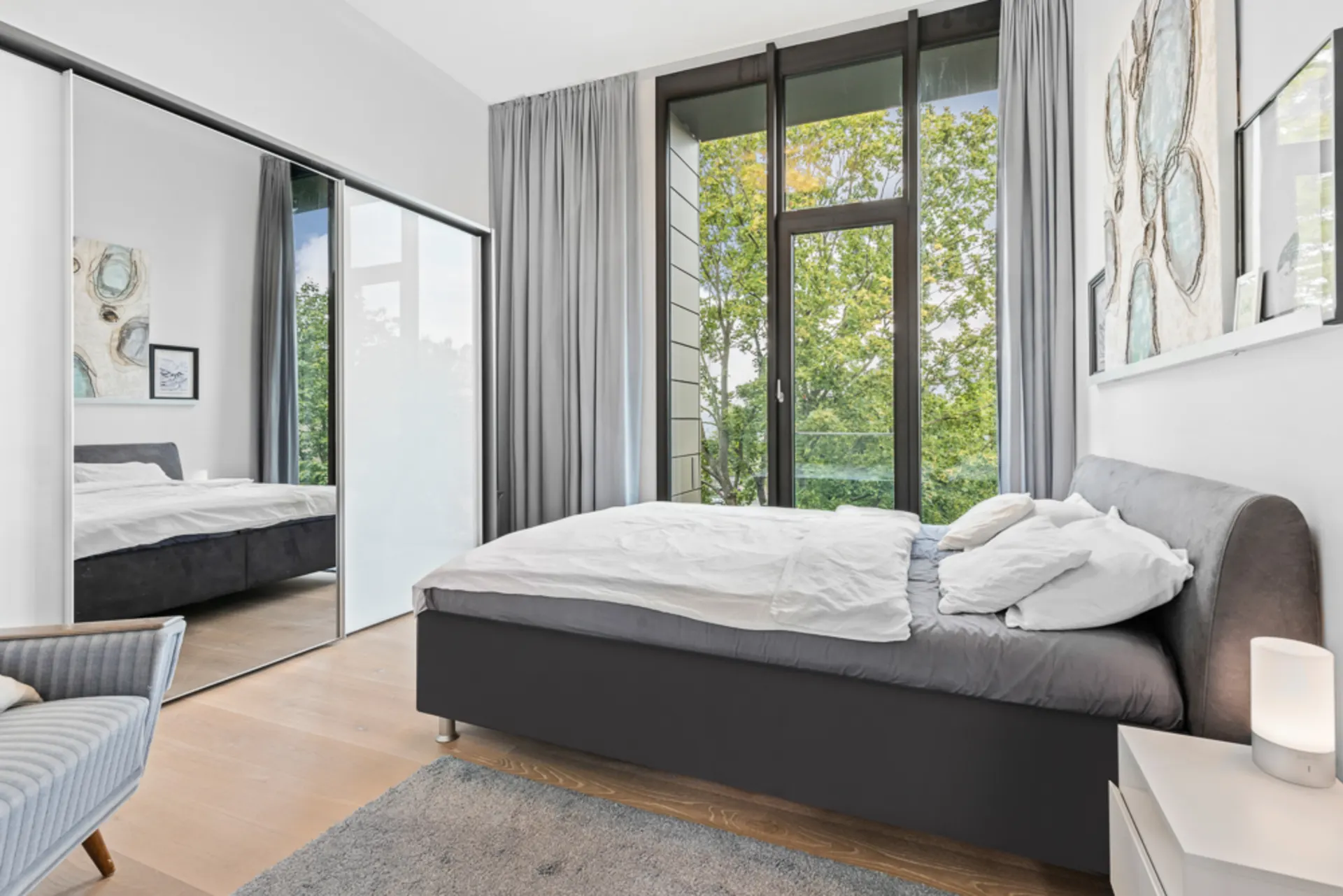 Modernes Schlafzimmer mit großem Bett, Spiegelkleiderschrank und bodentiefen Fenstern mit Blick ins Grüne.