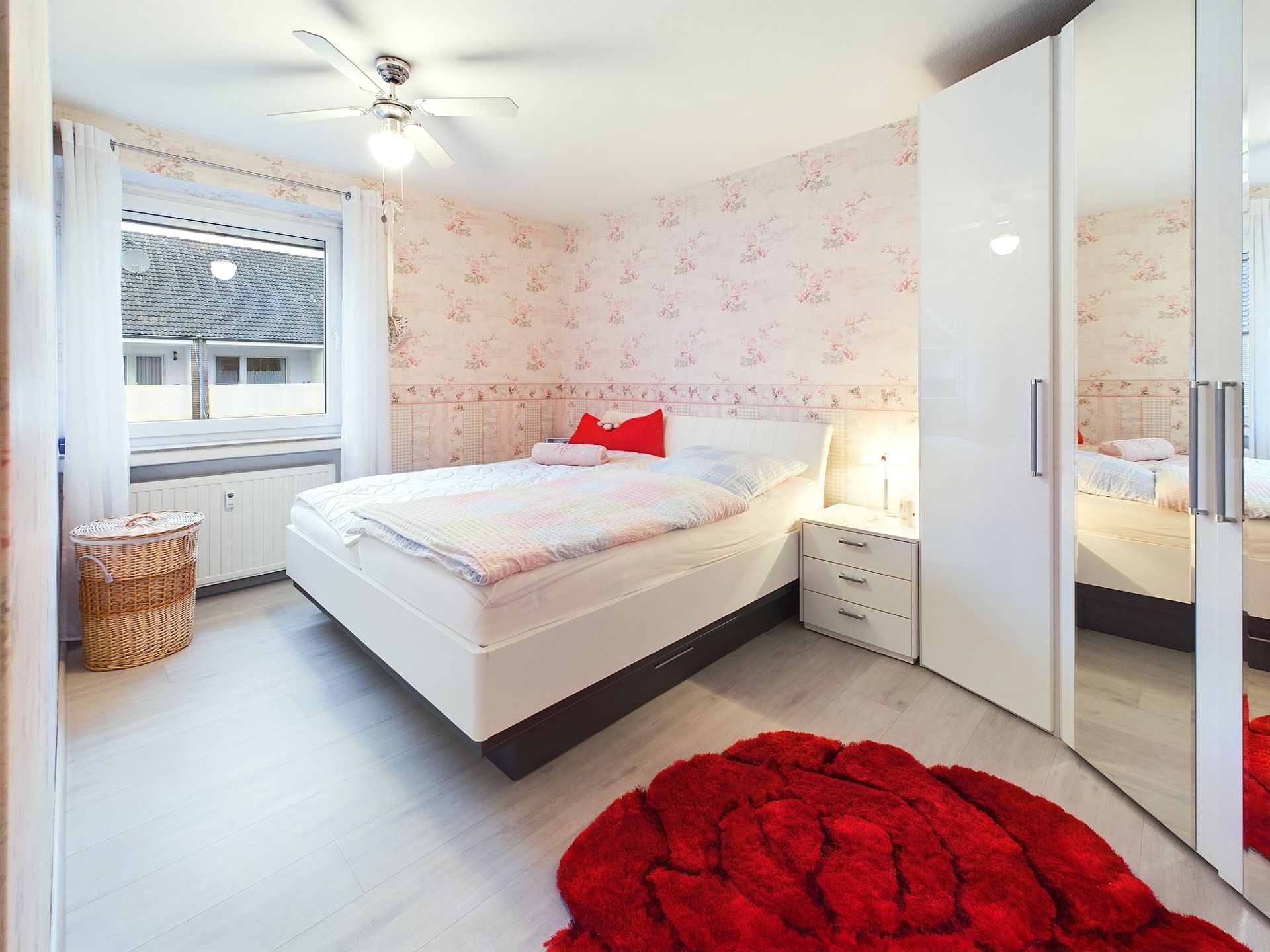 Helles Schlafzimmer mit Doppelbett, rotem Teppich, Kleiderschrank und Fenster.