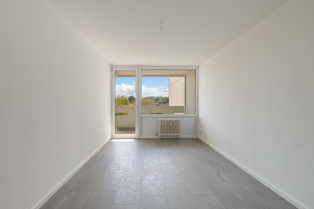 Leerer, heller Raum mit grauem Boden, weißem Heizkörper und Fenster zur Terrasse.