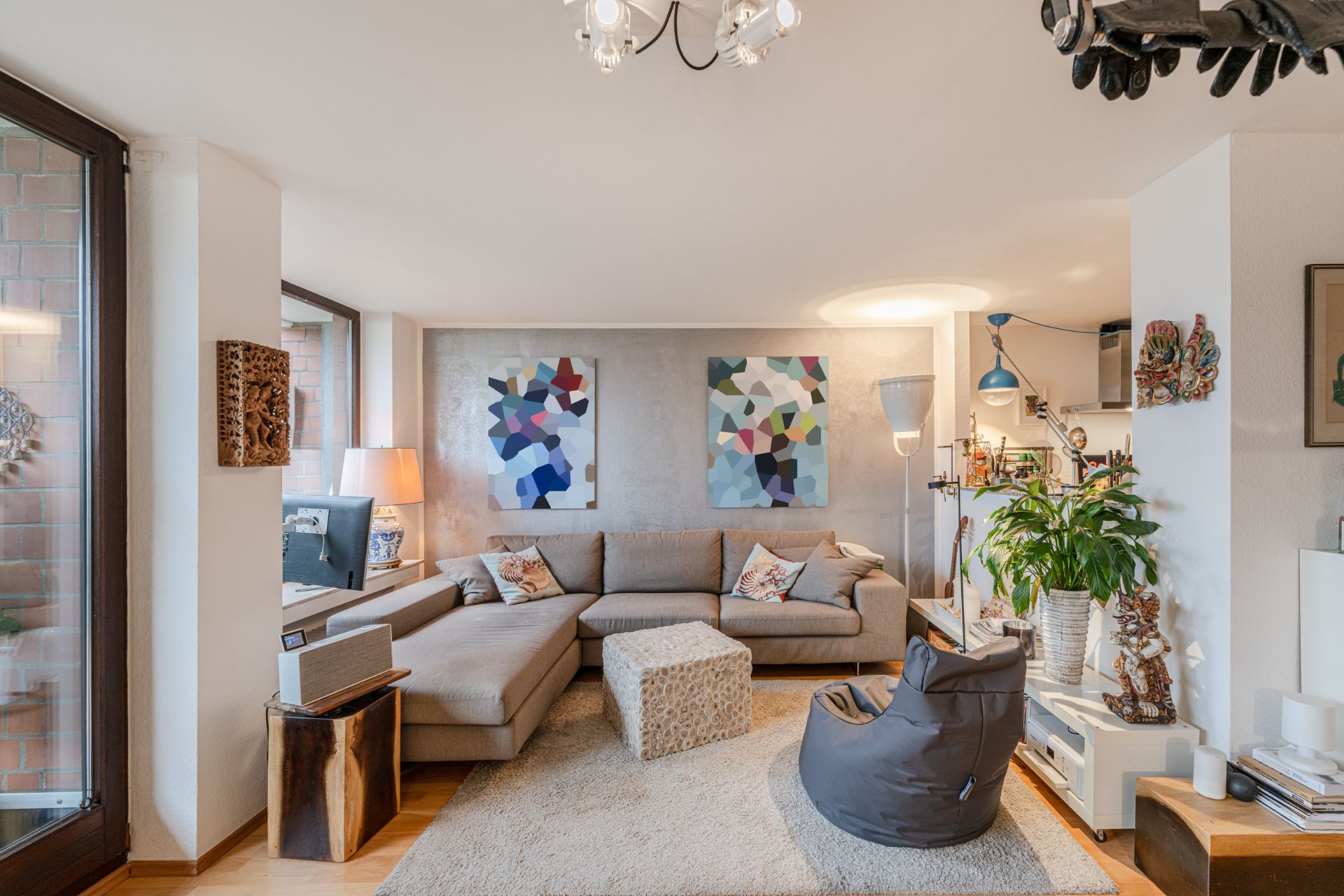 Wohnzimmer mit beigem Ecksofa, Teppich, Sessel und Pflanzen, bunte Bilder an der Wand.