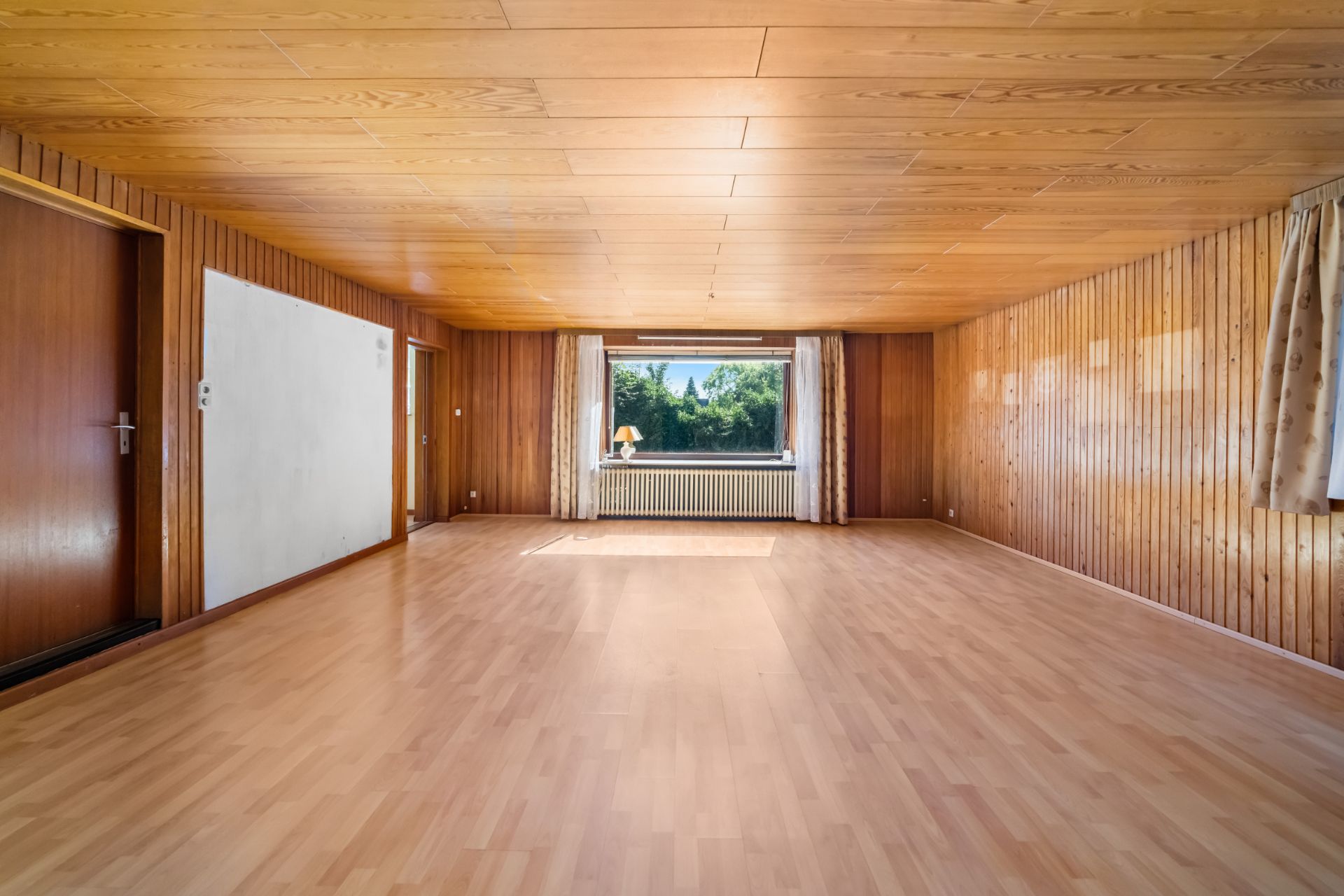 Leeres Wohnzimmer mit Holzvertäfelung, großem Fenster und Parkettboden.