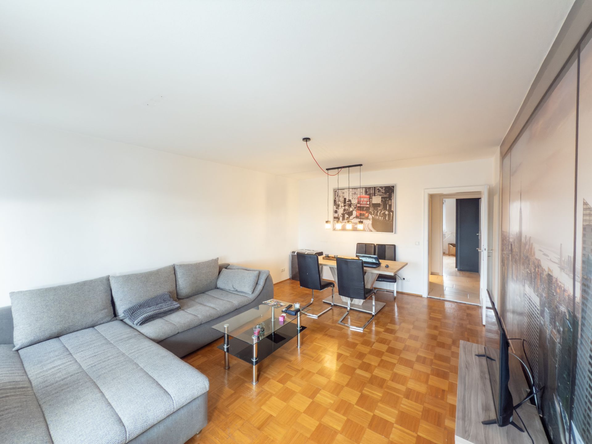 Helles Wohnzimmer mit grauem Ecksofa, Glastisch, Esstisch und Fernseher auf Parkettboden.