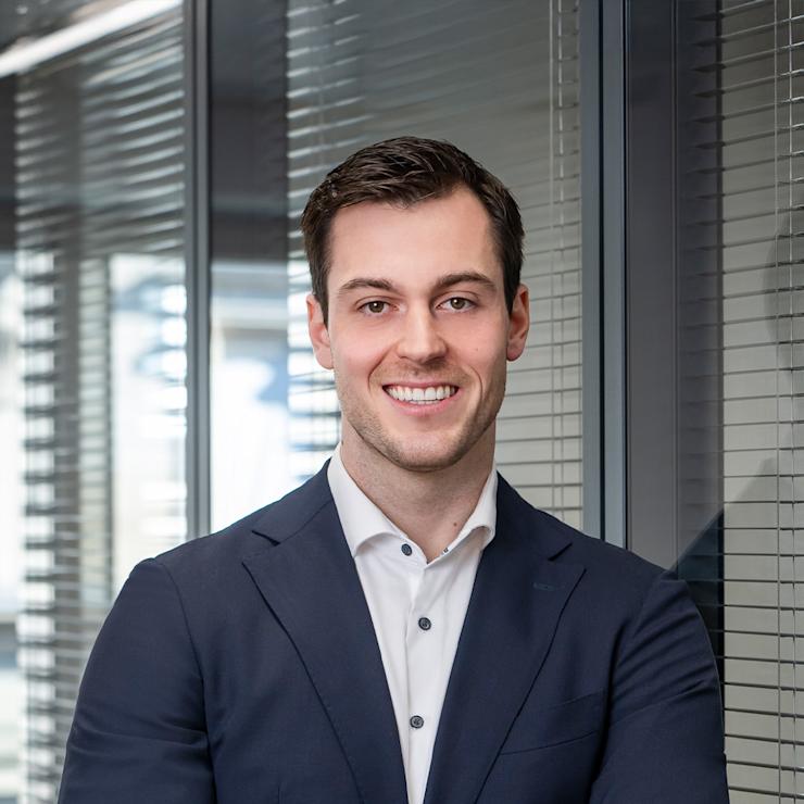 Mann in Anzug steht mit verschränkten Armen vor Glaswand mit Jalousien.