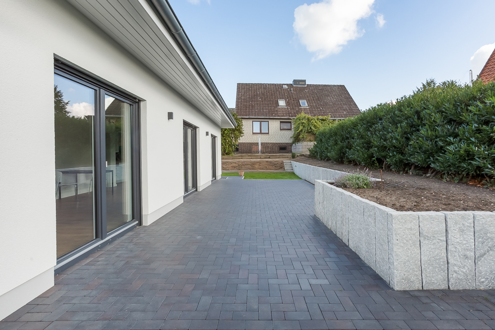 Moderne Terrasse mit Pflastersteinen, angrenzendem Gartenbeet und Blick auf ein Wohnhaus.