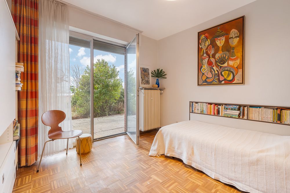 Helles Schlafzimmer mit Einzelbett, Bücherregal, Stuhl, offener Tür und großem Gemälde an der Wand.