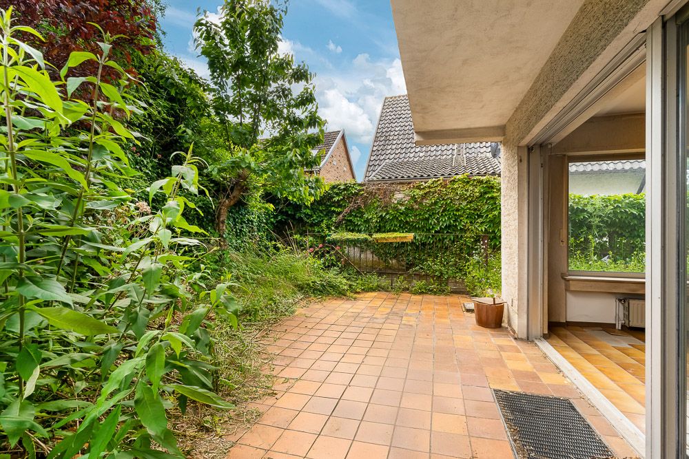Gepflasterte Terrasse mit offenen Türen zum Wohnbereich, umgeben von viel Grün und Pflanzen.