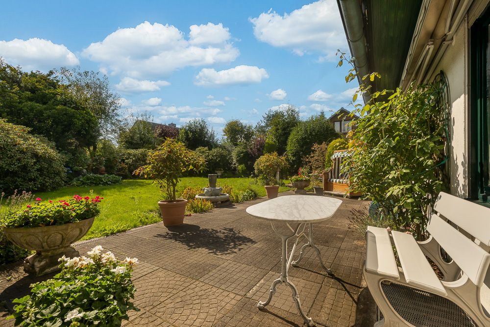 Sonnige Terrasse mit Tisch, Bank, Blumentöpfen und Blick auf einen grünen Garten.