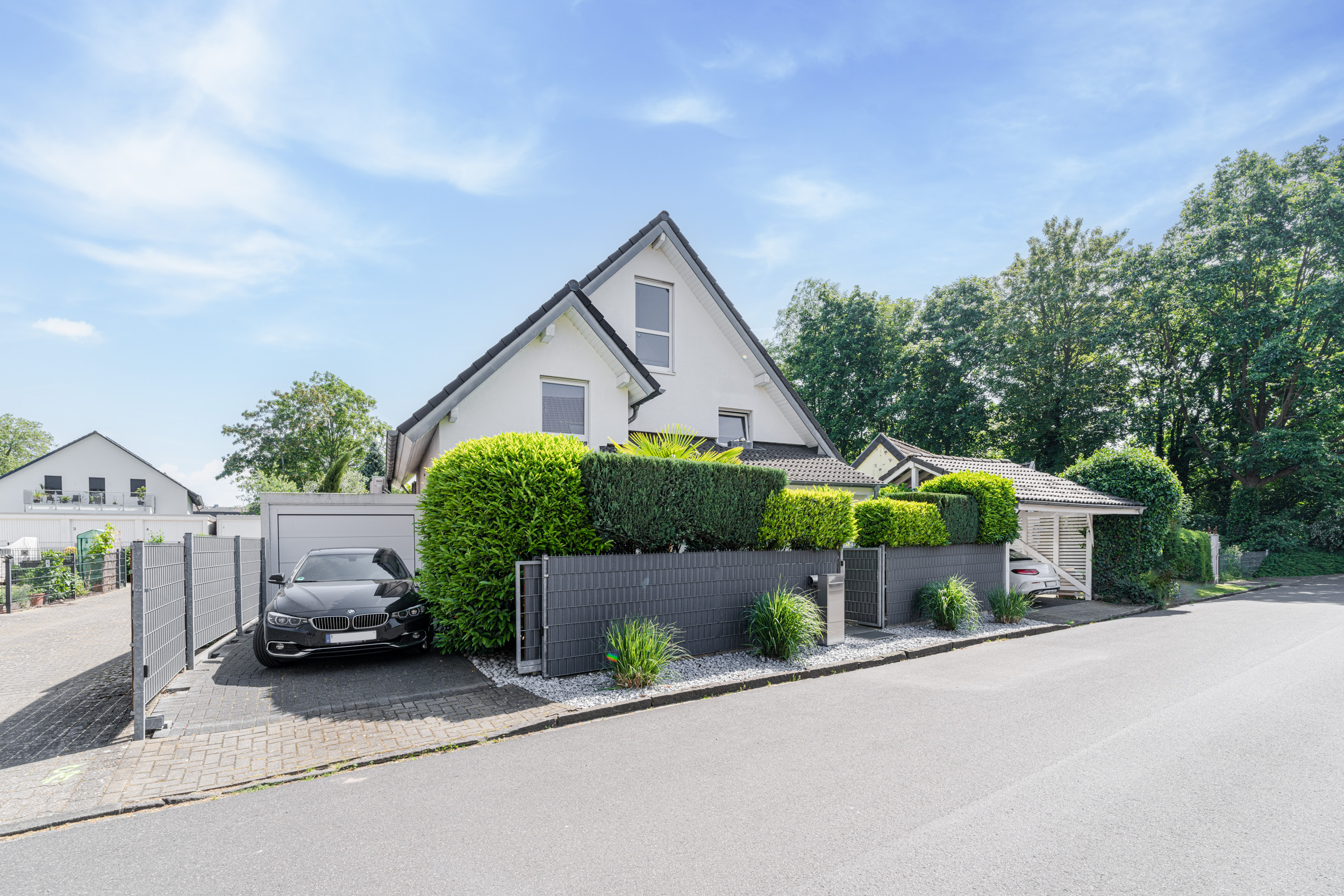 Modernes Einfamilienhaus mit grauem Zaun, Einfahrt und geparktem Auto vor gepflegten Hecken.