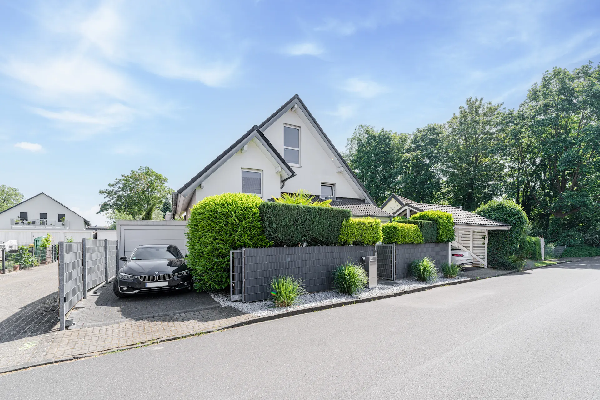 Modernes Einfamilienhaus mit grauem Zaun, Einfahrt und geparktem Auto vor gepflegten Hecken.