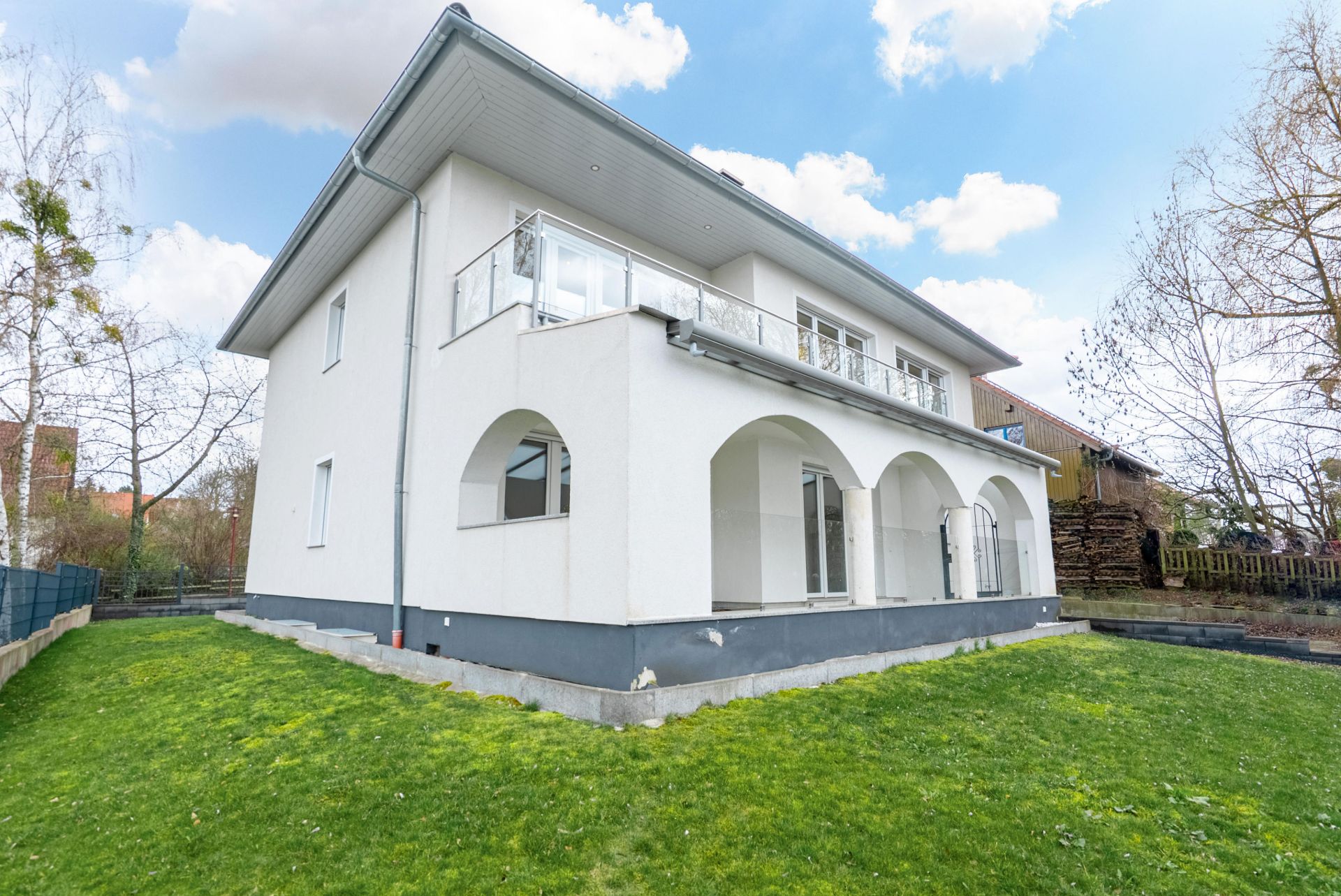 Zweistöckiges weißes Haus mit Balkon, Bögen und Garten, umgeben von Bäumen.