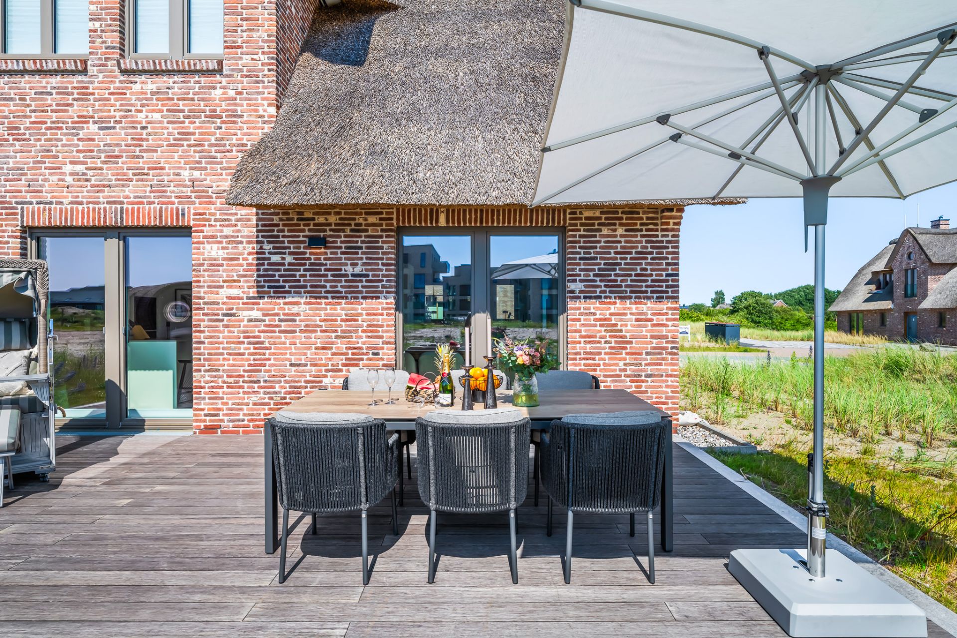 Terrasse mit großem Esstisch, Stühlen, Sonnenschirm und Backsteinhaus im Hintergrund.