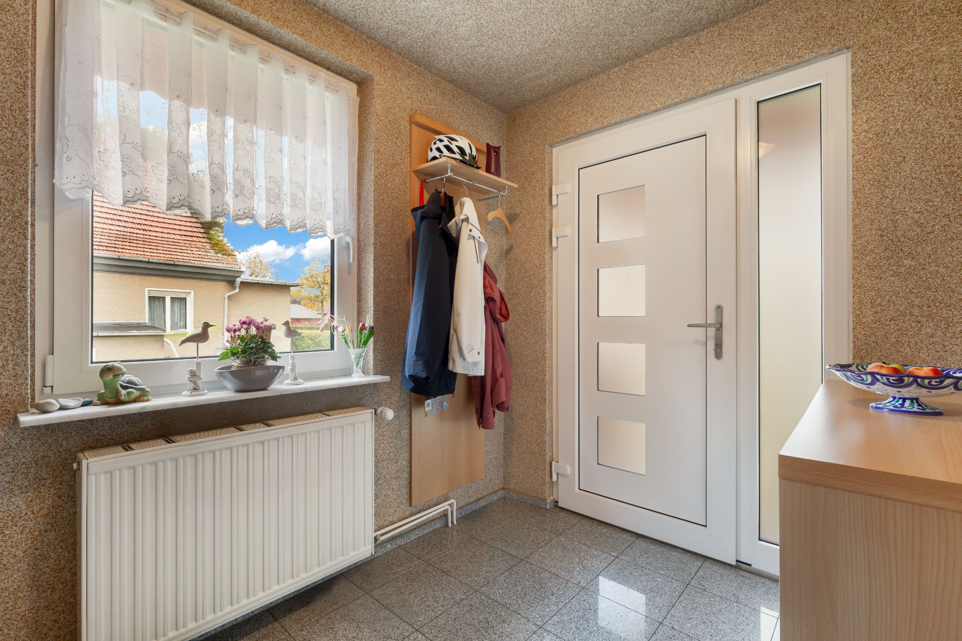 Heller Eingangsbereich mit weißer Tür, Garderobe, Fenster und Kommode mit Schale.