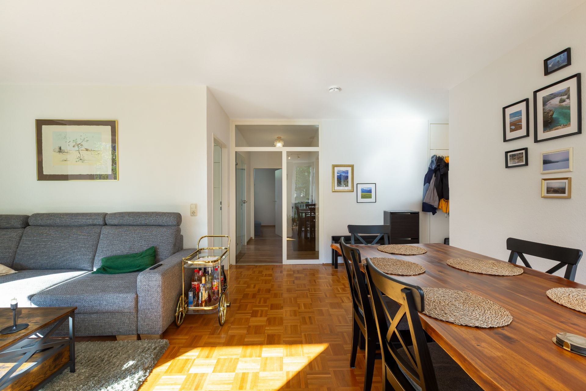 Helles Wohnzimmer mit grauem Sofa, Esstisch, Holzboden und Bildern an der Wand.