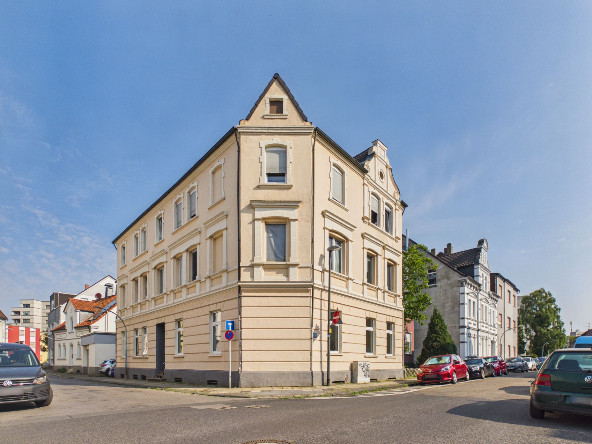 Eckhaus mit hellem Anstrich an einer Straßenkreuzung, mehrere geparkte Autos sichtbar.
