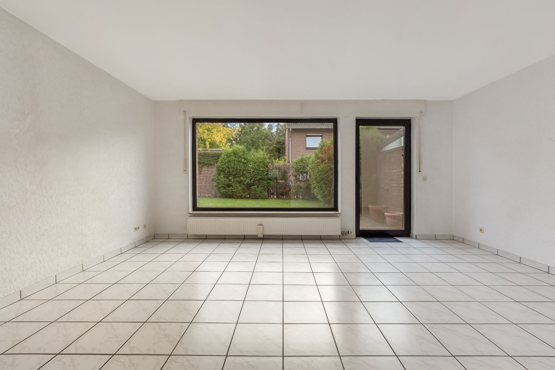Leeres Zimmer mit weißen Fliesen, großem Fenster und Glastür zum Garten.