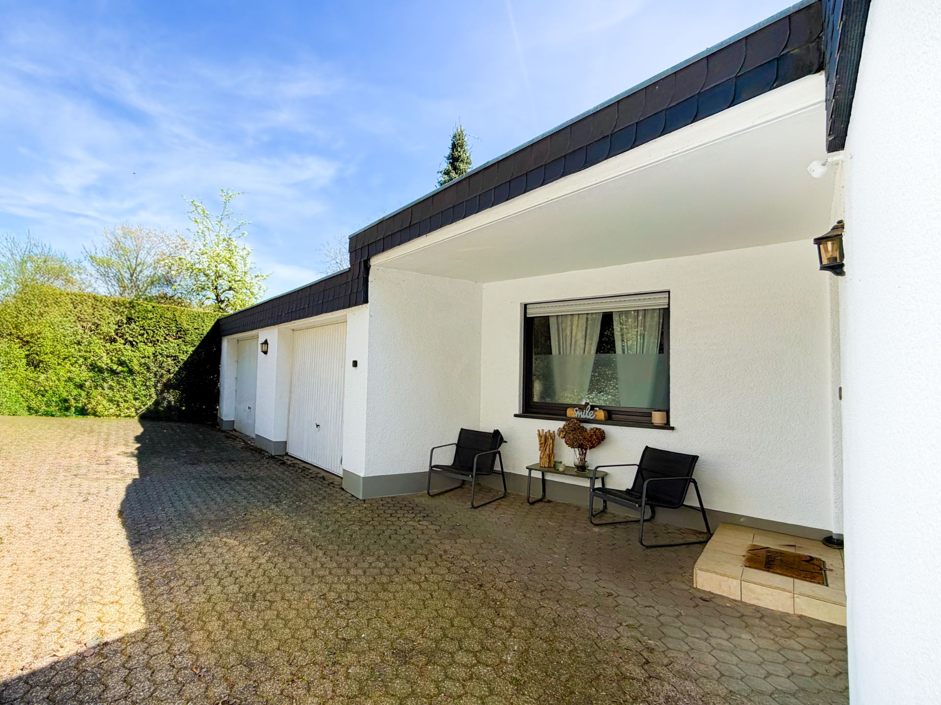 Überdachter Hauseingang mit zwei Stühlen, Tisch und Garage auf gepflastertem Hof.
