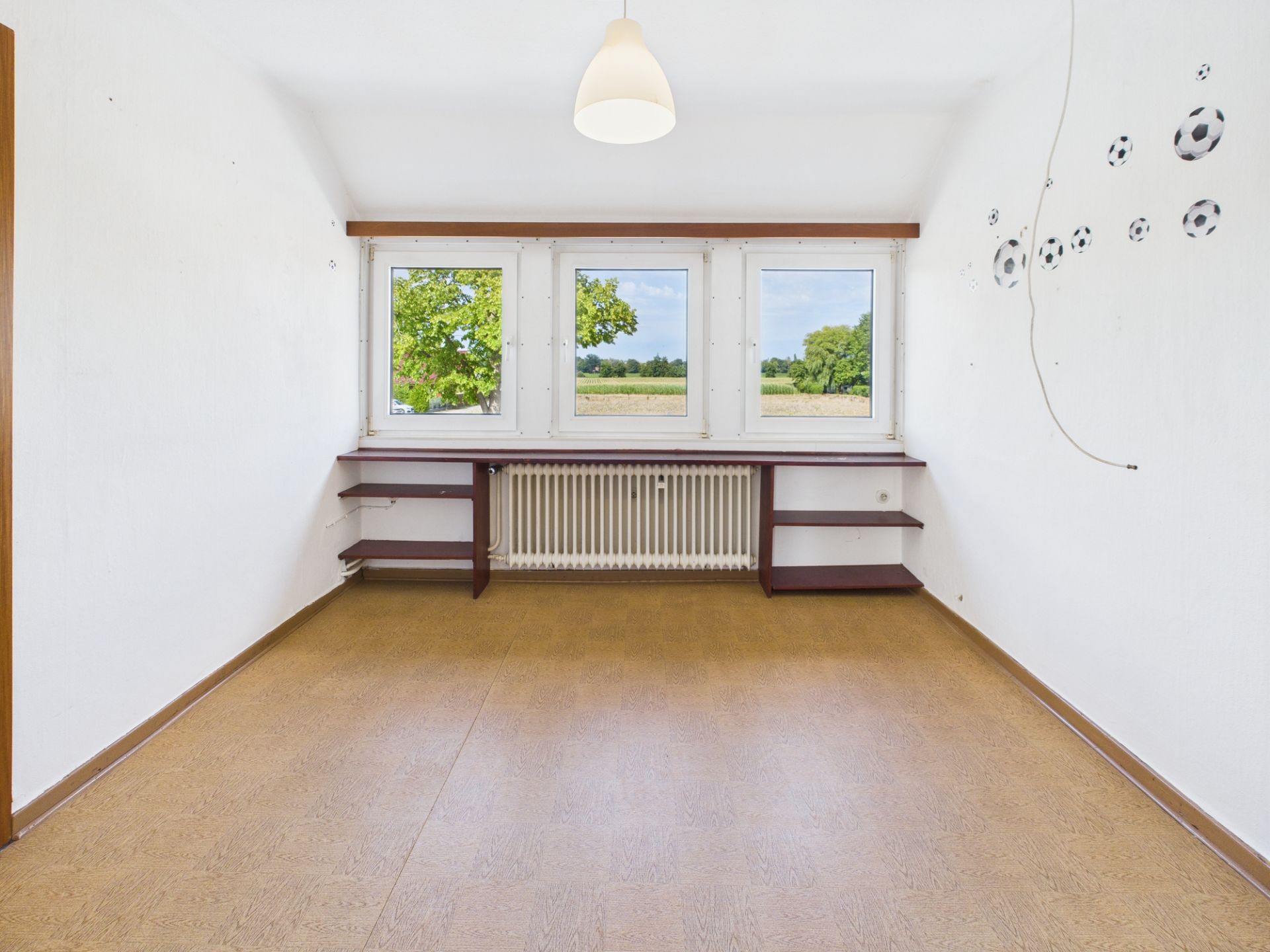 Leerer Raum mit drei Fenstern, Wandregalen und Fußballaufklebern an der weißen Wand.