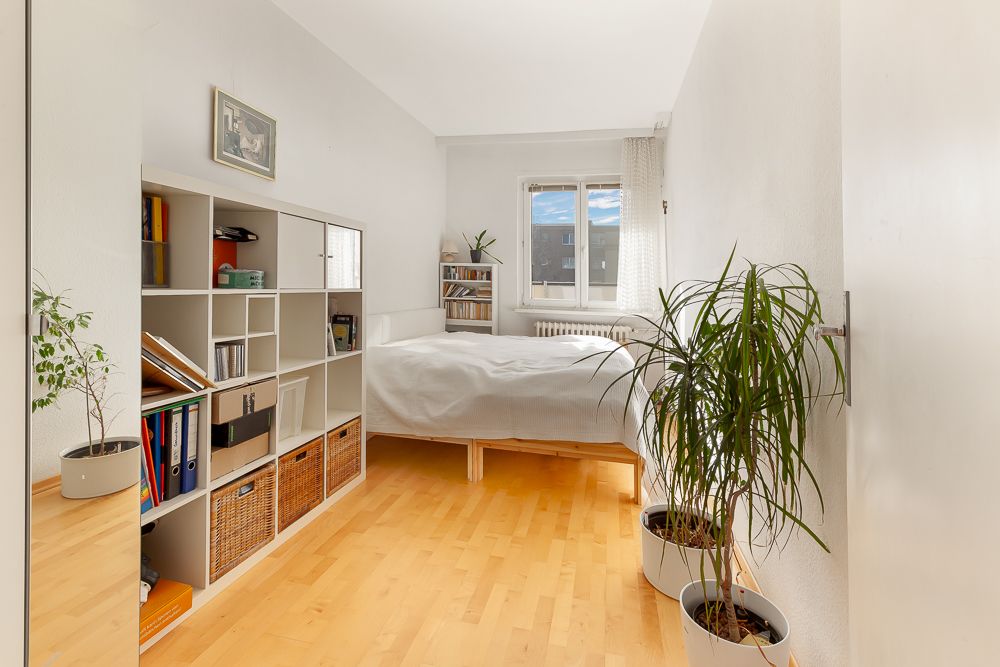 Helles Schlafzimmer mit Holzboden, Regal, Bett, Pflanzen und Fenster mit Blick nach draußen.