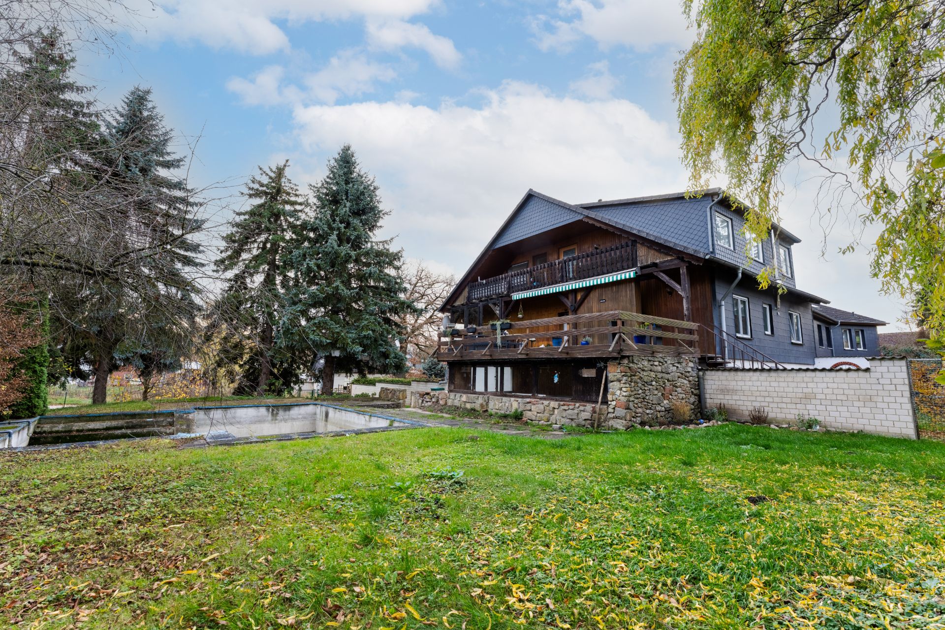 Großes Haus mit Holzveranda, Steinmauer und Garten mit leerem Pool, Bäume im Hintergrund.