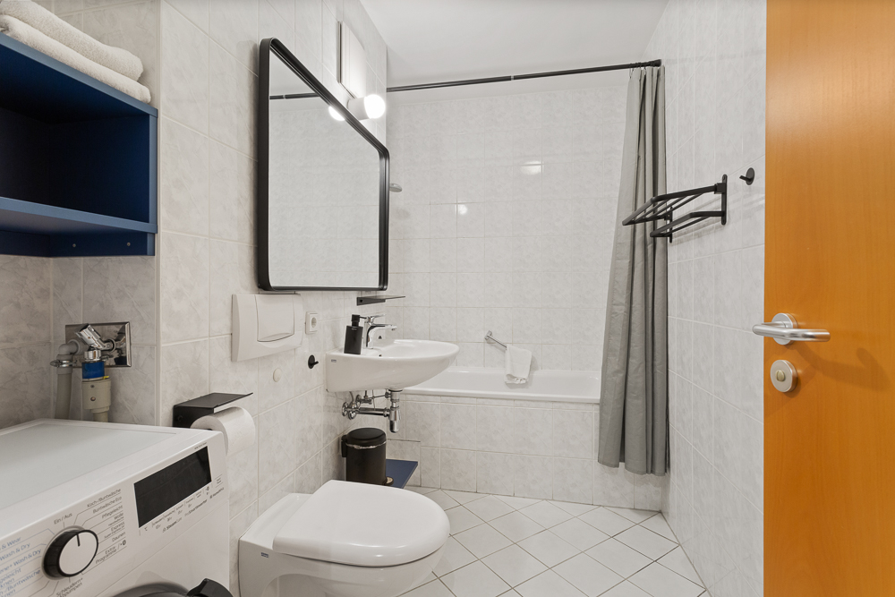Modernes Badezimmer mit Badewanne, Waschbecken, WC, Spiegel, Waschmaschine und Handtüchern.