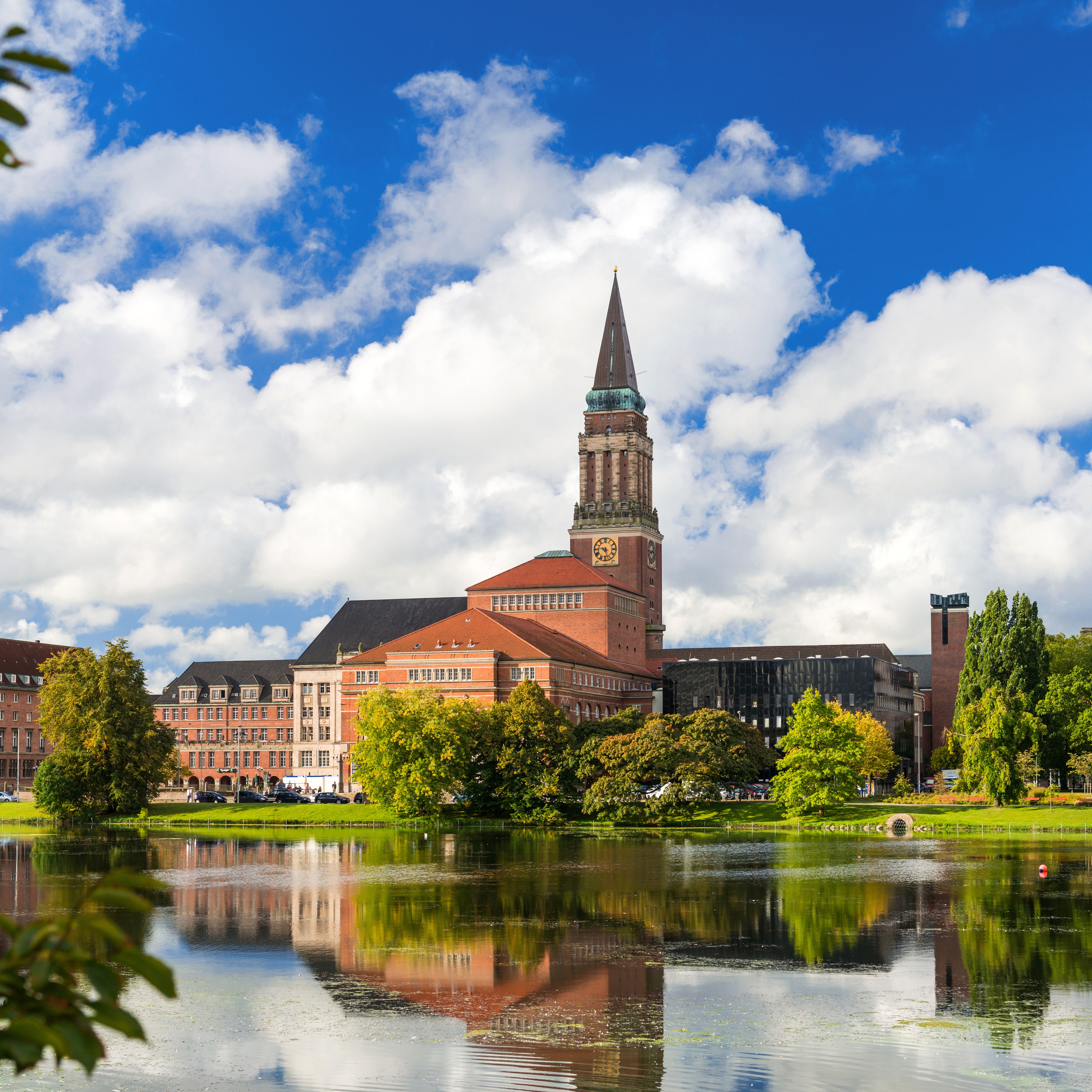 Rotes Backsteingebäude mit Turm an einem Seeufer, umgeben von Bäumen.
