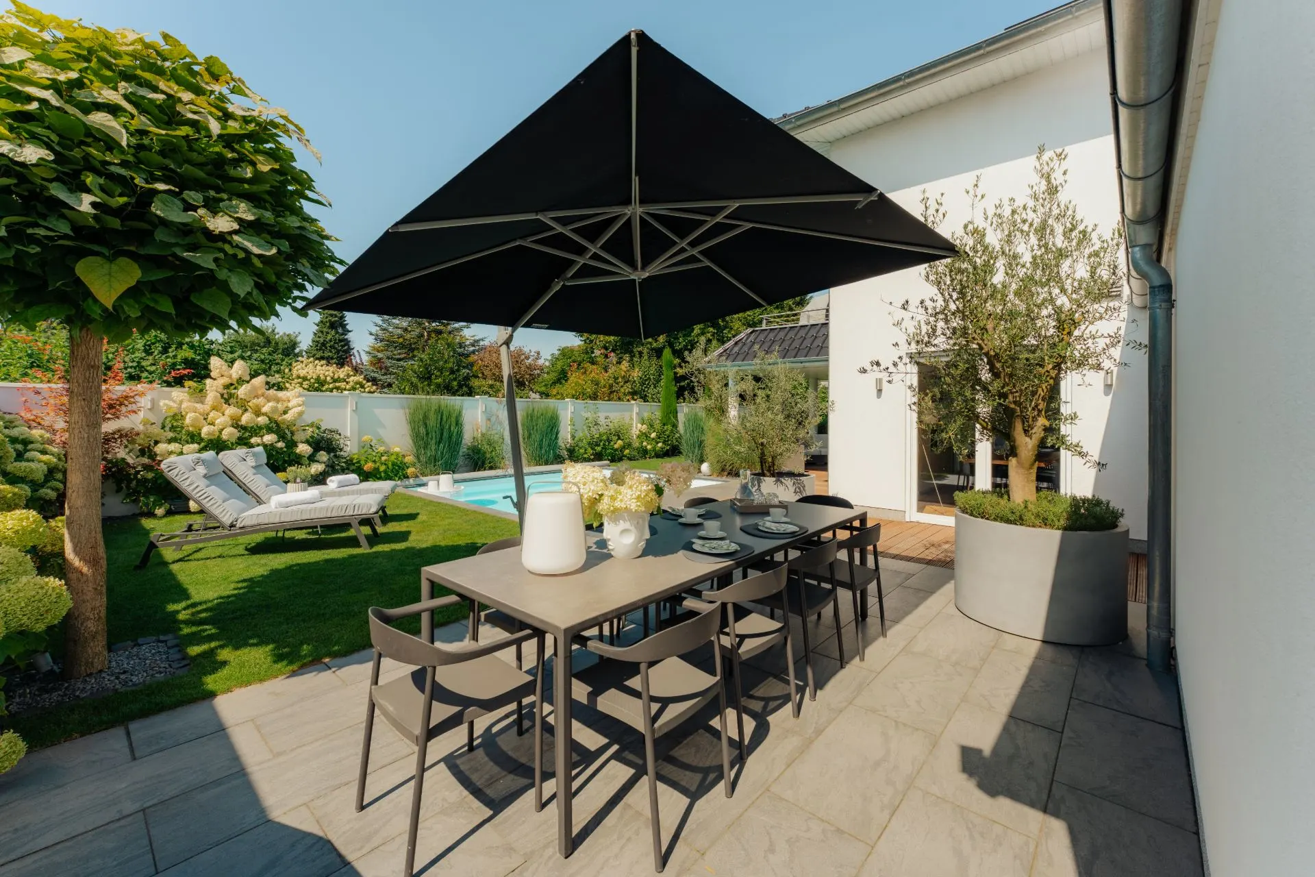 Moderne Terrasse mit Esstisch, Stühlen, großem Sonnenschirm und Blick auf Garten mit Pool.
