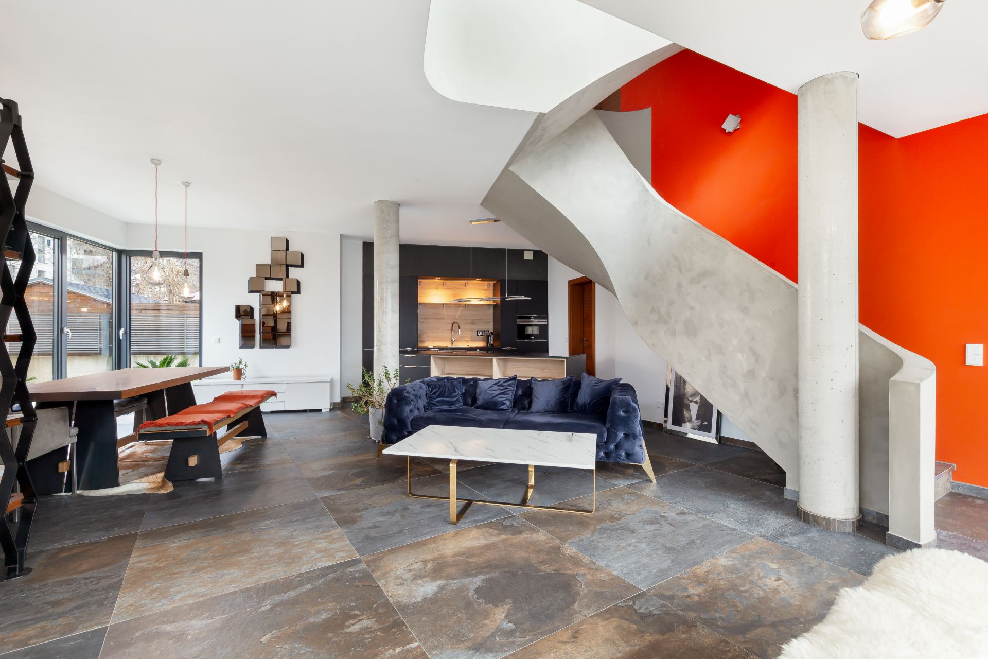Modernes Wohnzimmer mit grauer Treppe, blauer Couch und Esstisch auf großen Fliesen.