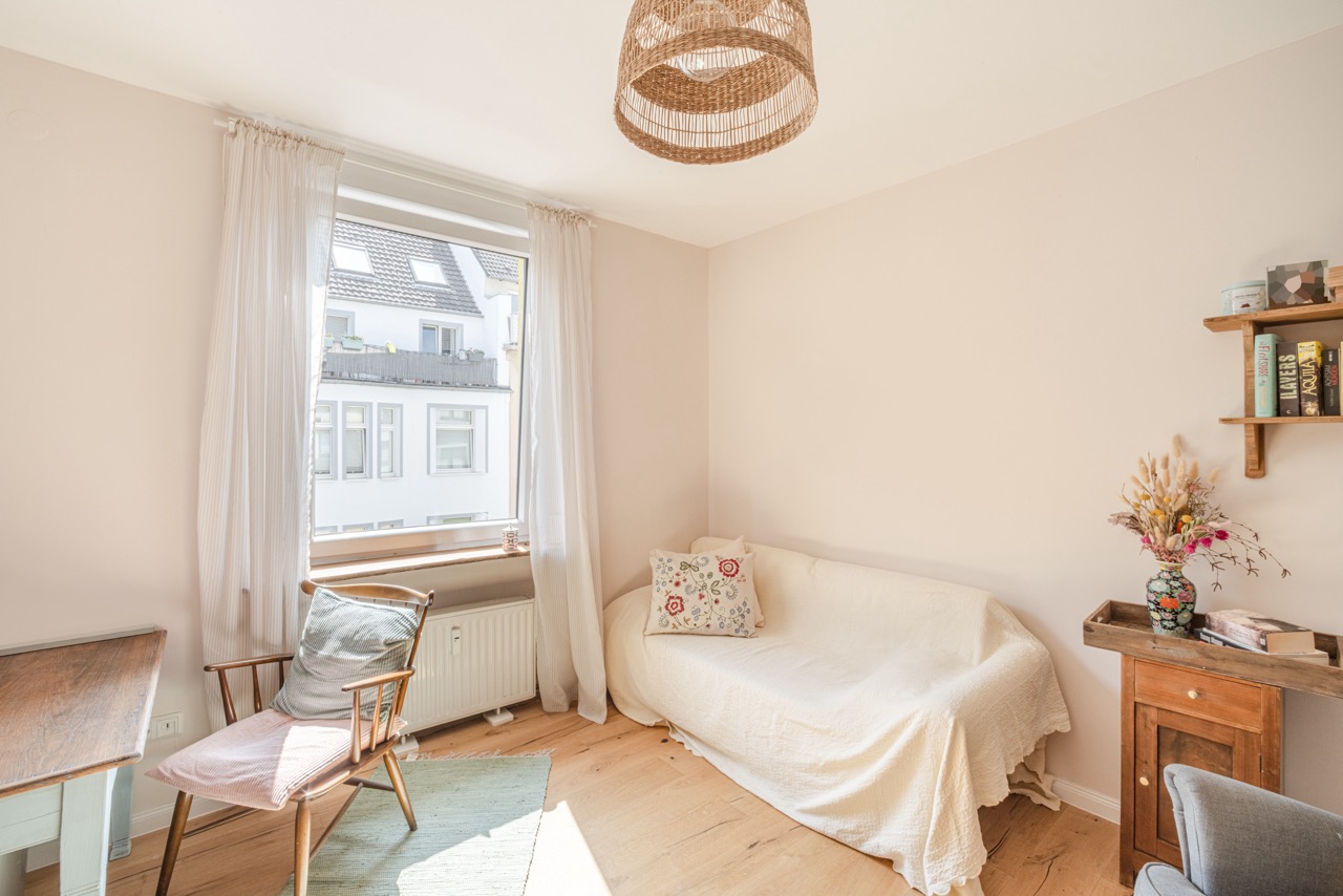 Helles Wohnzimmer mit Sofa, Holzstuhl, Fenster, Bücherregal und Blumenvase auf kleinem Schrank.