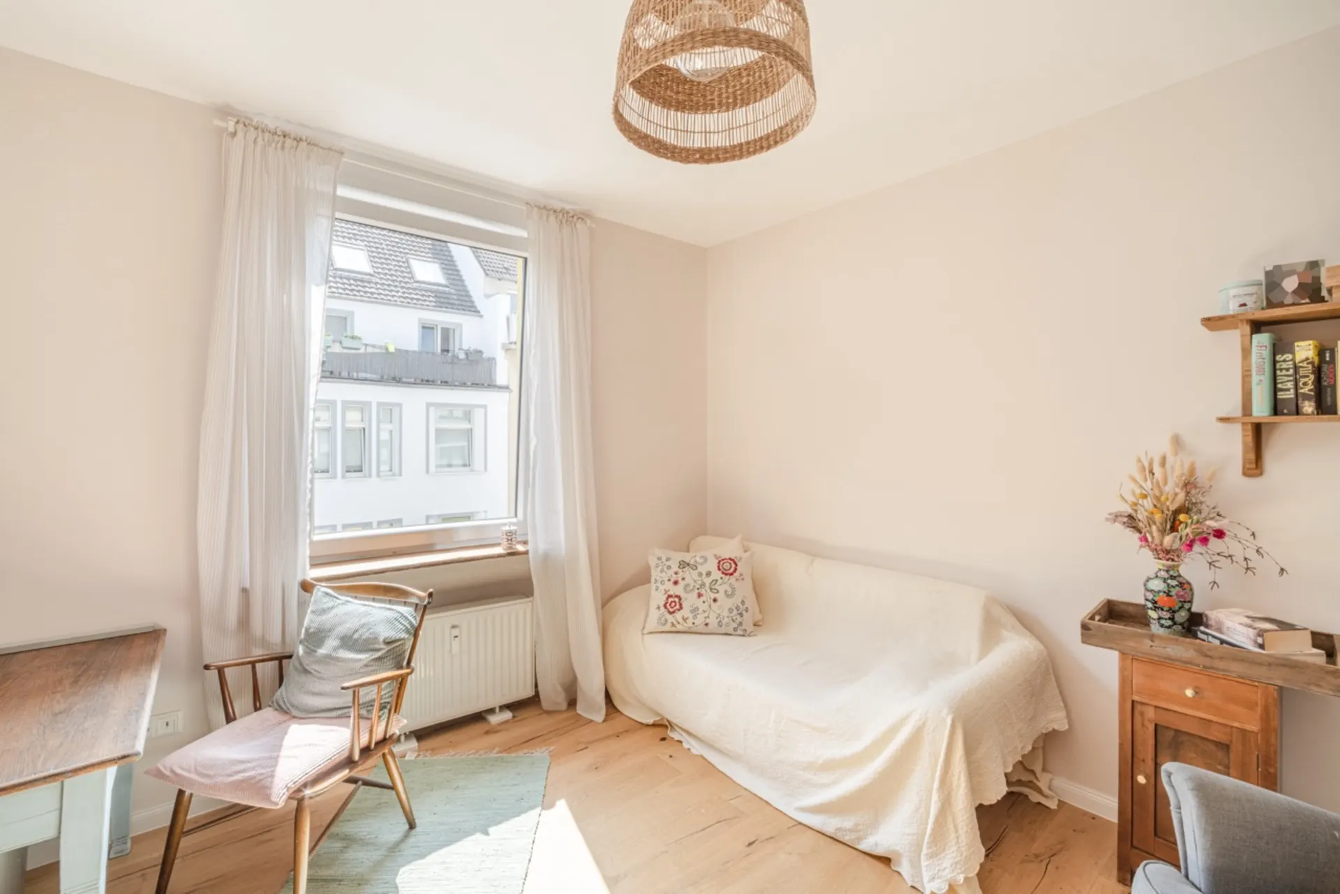 Helles Wohnzimmer mit Sofa, Holzstuhl, Fenster, Bücherregal und Blumenvase auf kleinem Schrank.