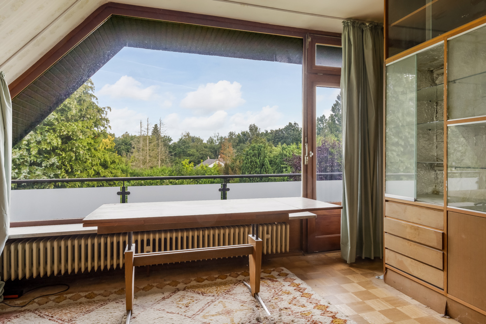 Helles Zimmer mit großem Fenster, Holztisch, Teppich und Blick auf Bäume im Garten.