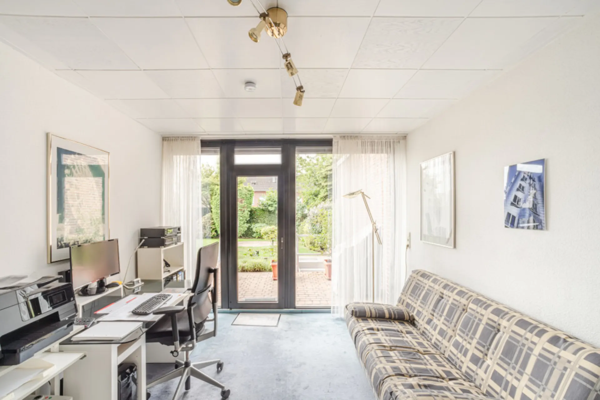 Helles Arbeitszimmer mit Schreibtisch, Sofa und Zugang zum Garten durch eine Glastür.