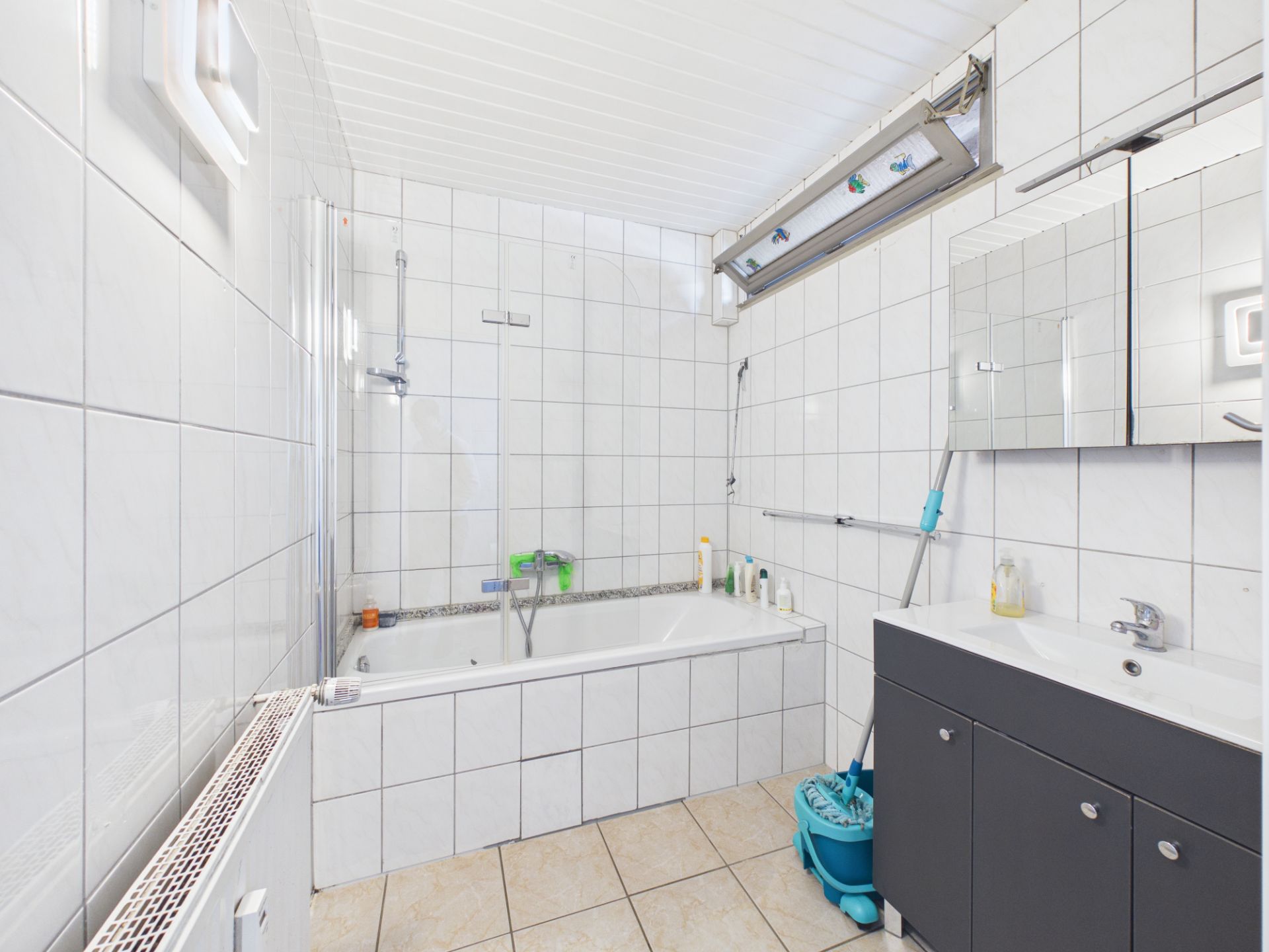 Helles Badezimmer mit Badewanne, Waschbecken, Spiegelschrank und Reinigungsutensilien.