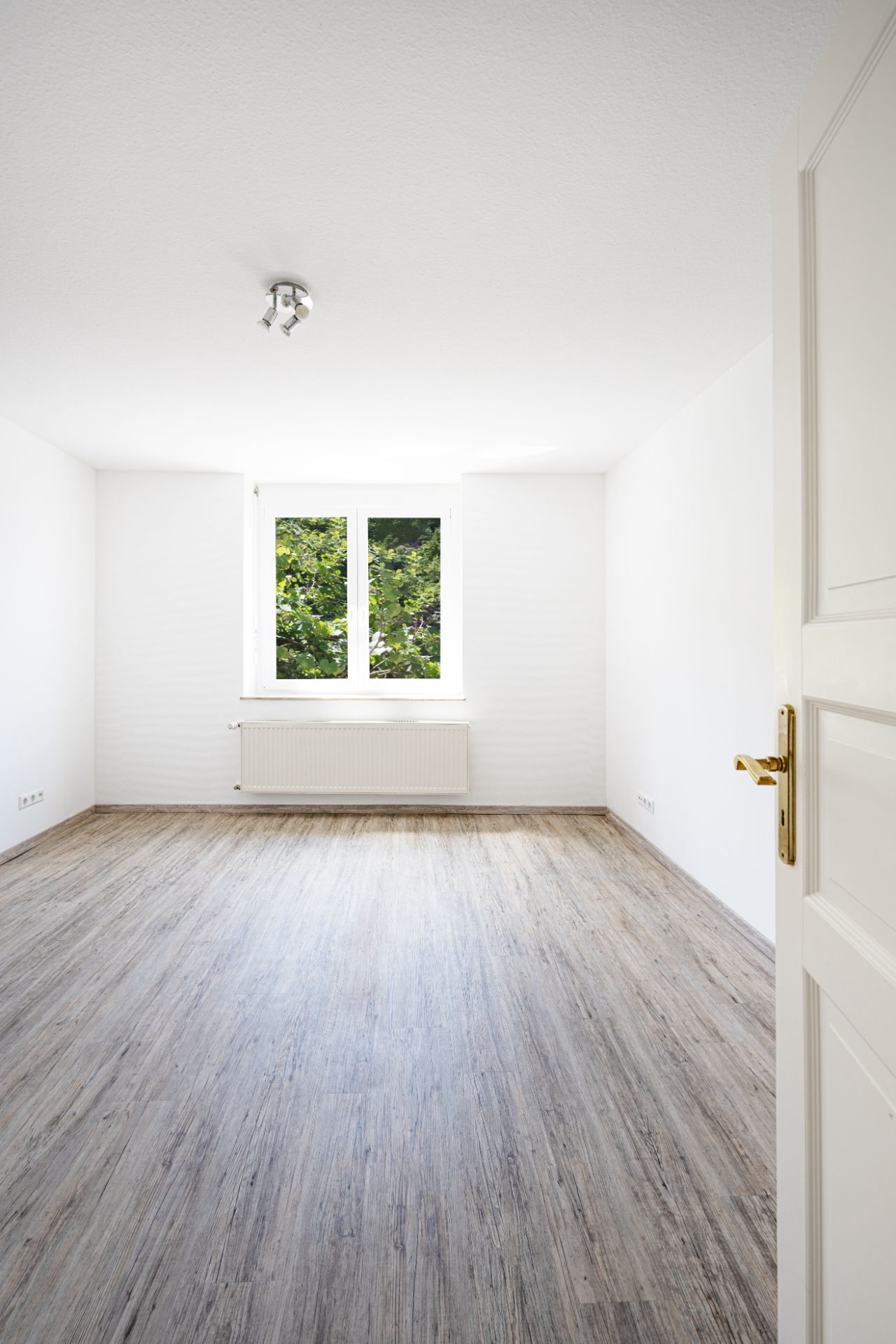 Leeres, helles Zimmer mit Holzfußboden, weißem Fenster und Heizkörper unter dem Fenster.