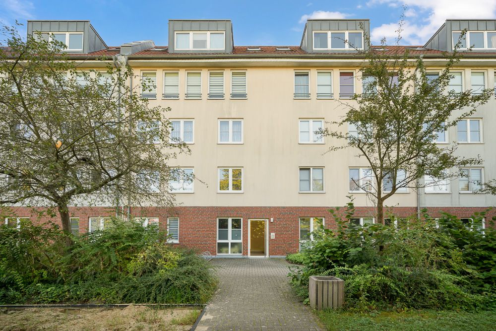 Vierstöckiges Wohnhaus mit Flachdach und Bäumen davor, Gehweg führt zum Eingang.