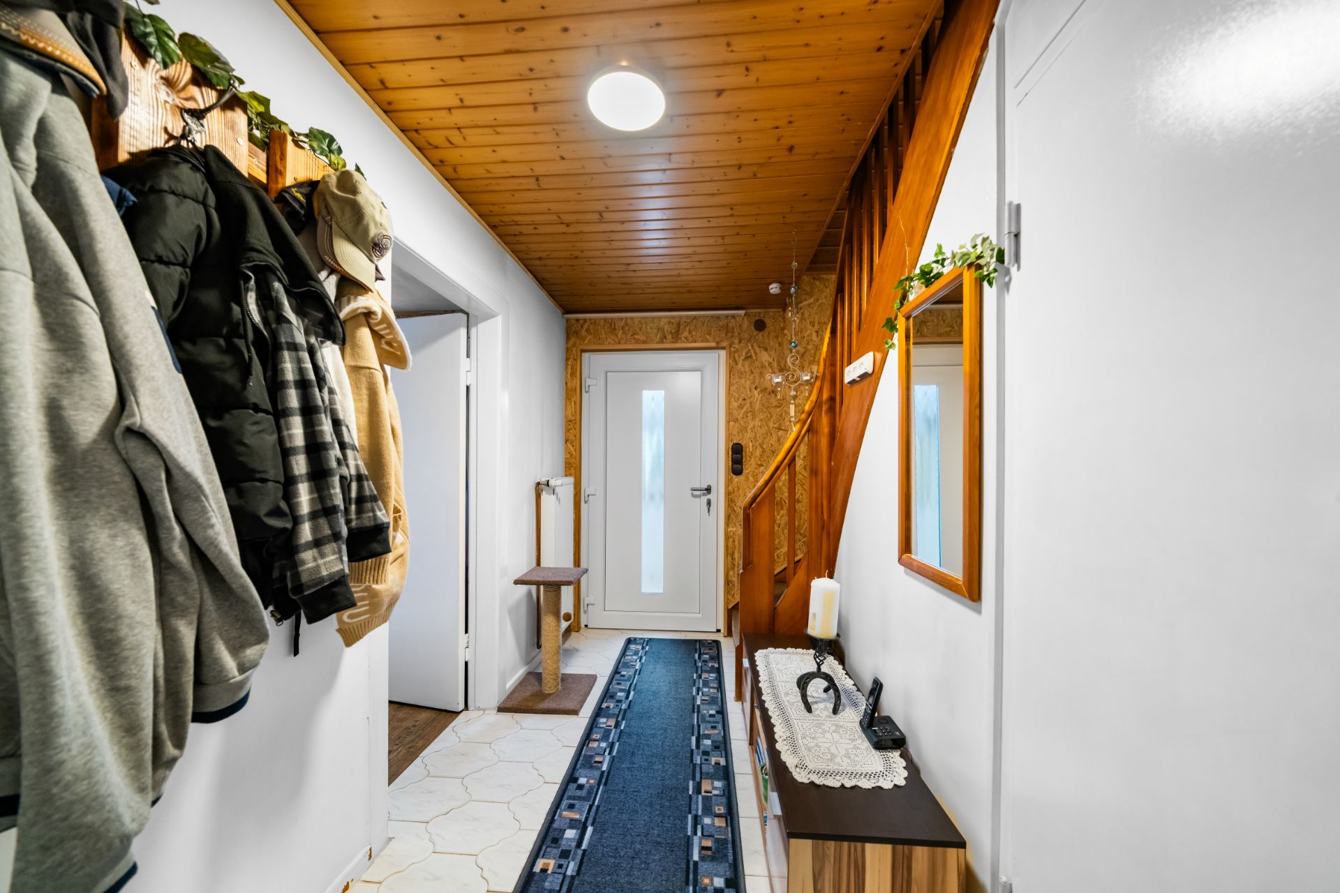 Heller Flur mit Holztreppe, Garderobe, Teppichläufer und Spiegel an der Wand.