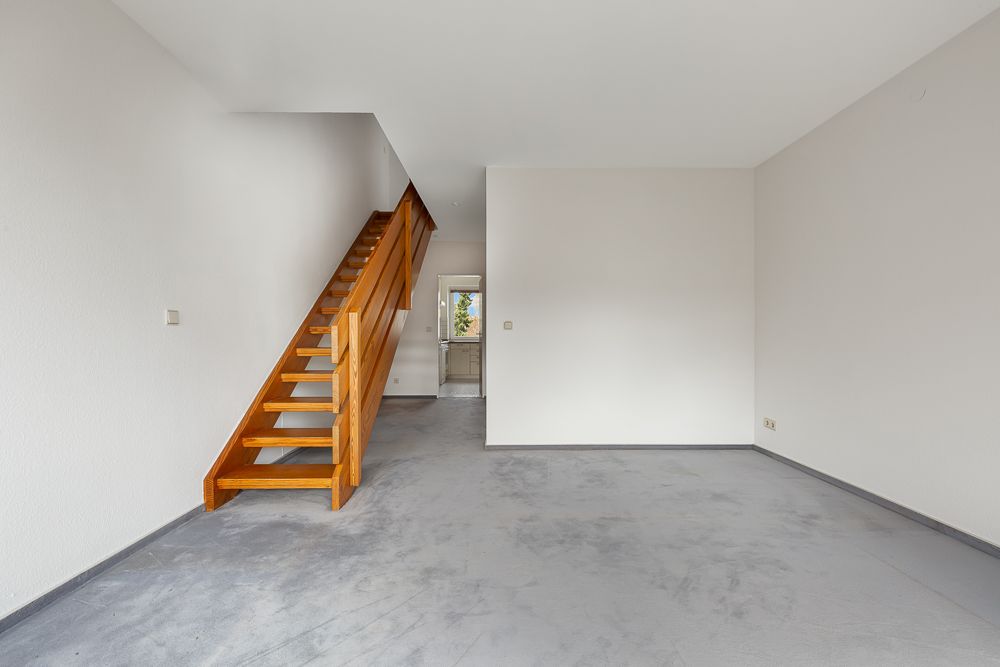 Leerer Raum mit Holztreppe und Durchgang zur Küche, Wände und Boden schlicht gehalten.
