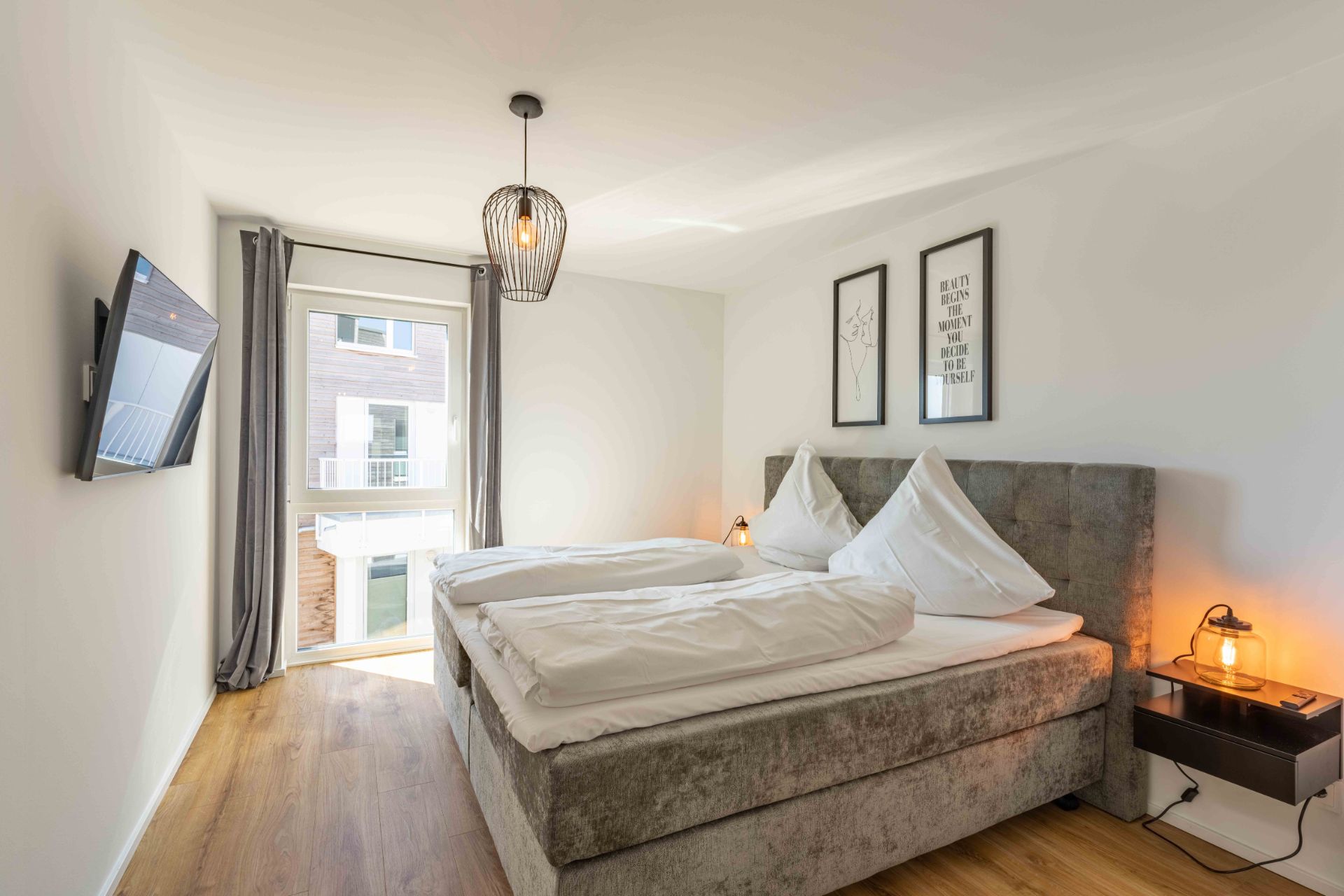Helles Schlafzimmer mit Doppelbett, Wandbildern, TV, Nachttisch und Fenster mit Vorhängen.